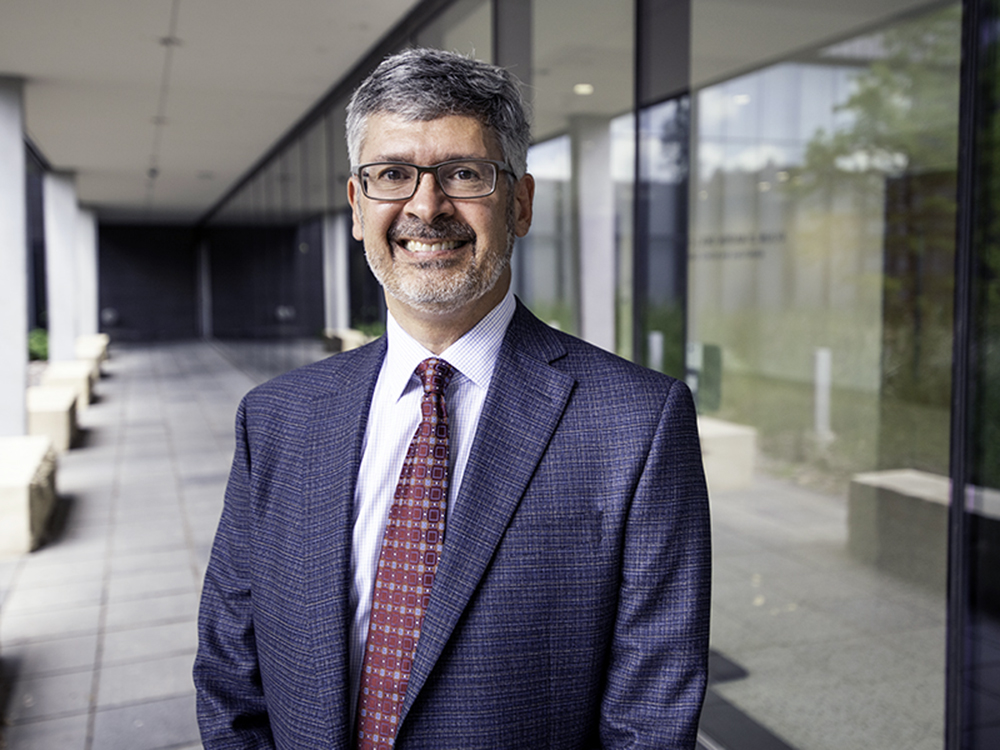 Roger Bonnecaze stands outside the EER building on the UT Austin campus. A tall, slim man, he's wearing a gray suit with a red tie and dary eye glasses.