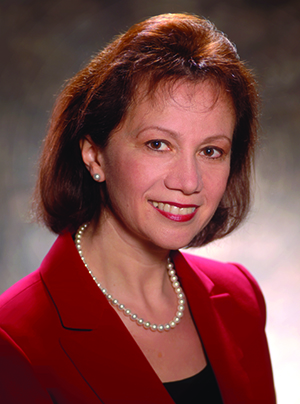 Bella Goren, a 1983 graduate of the Chemical Engineering Department at UT Austin, wearing a red suit, black top and pearl necklace sitting in front of a gray background.